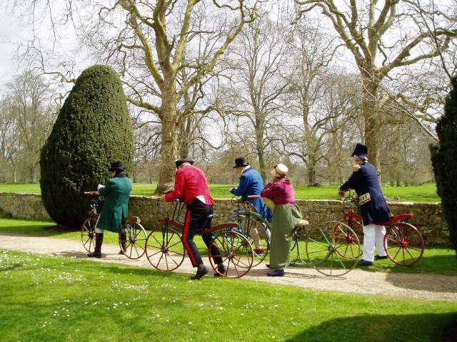 Men and women in Regency dress riding velocipedes on the grounds of Montacute House